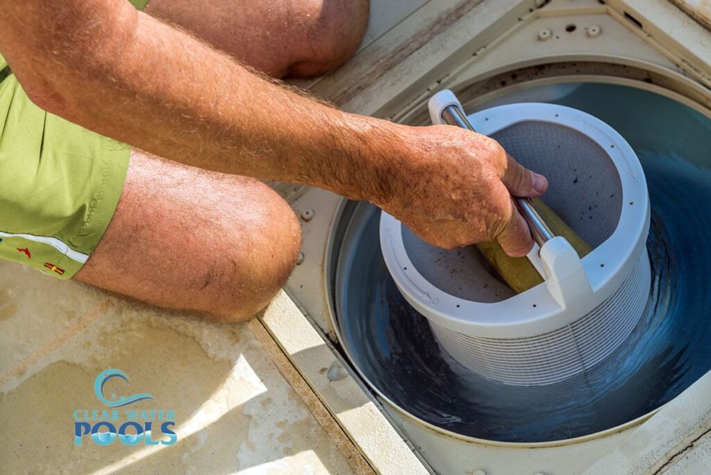 A person in green shorts removes a pool skimmer basket from a pool filter compartment, demonstrating proper pool maintenance, with a "Clear Water Pools" logo visible on the bottom left.