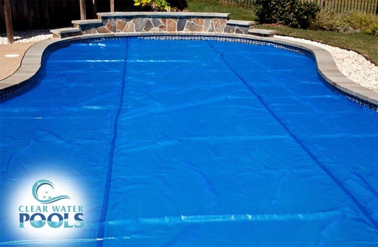 A backyard swimming pool covered with a blue pool cover is surrounded by landscaping and a stone wall. The "Clear Water Pools" logo appears in the lower left corner.