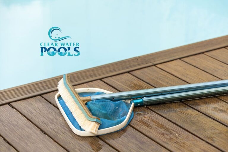 A pool brush and skimmer net rest on a wooden deck next to a clear swimming pool, illustrating how to clean a pool, with the "Clear Water Pools" logo in the background.