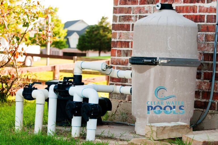 A pool filtration system with PVC pipes and a large filter tank labeled "Clear Water Pools" is installed next to a brick wall outdoors.