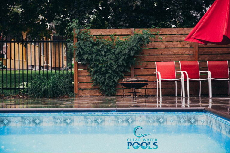 A backyard pool scene with red chairs, a red umbrella, wet pavement, a wooden fence, and greenery. The Clear Water Pools logo is visible in the pool, emphasizing proper pool water levels for a perfect swim experience.
