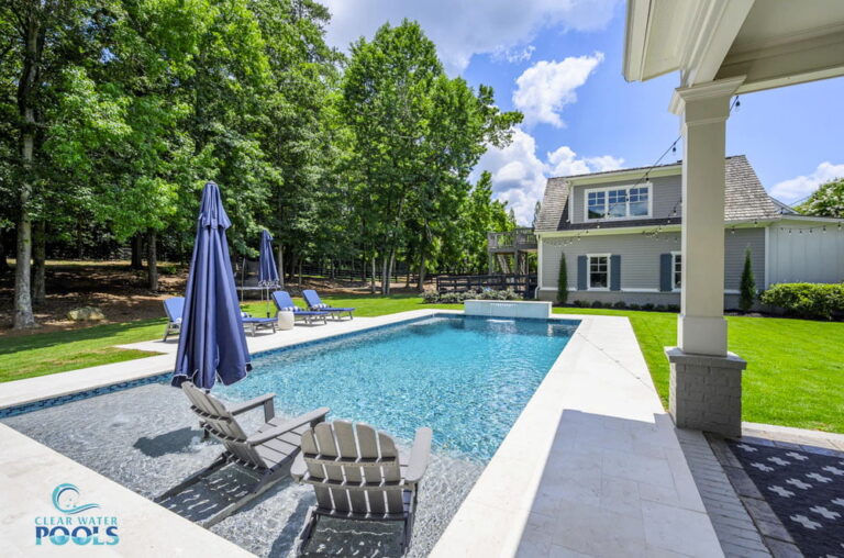 Rectangular gunite pool with clear water, surrounded by lounge chairs and umbrellas, sits beside a lush lawn and a two-story house on a sunny day.