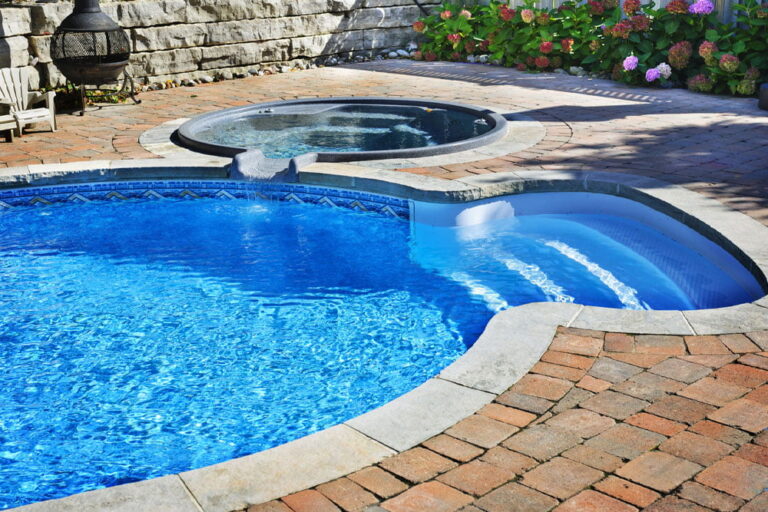 An outdoor pool with curved steps and an adjacent hot tub, surrounded by a brick patio, a stone wall, and blooming hydrangea bushes—perfect for relaxing in inviting hot tubs amid beautiful garden scenery.