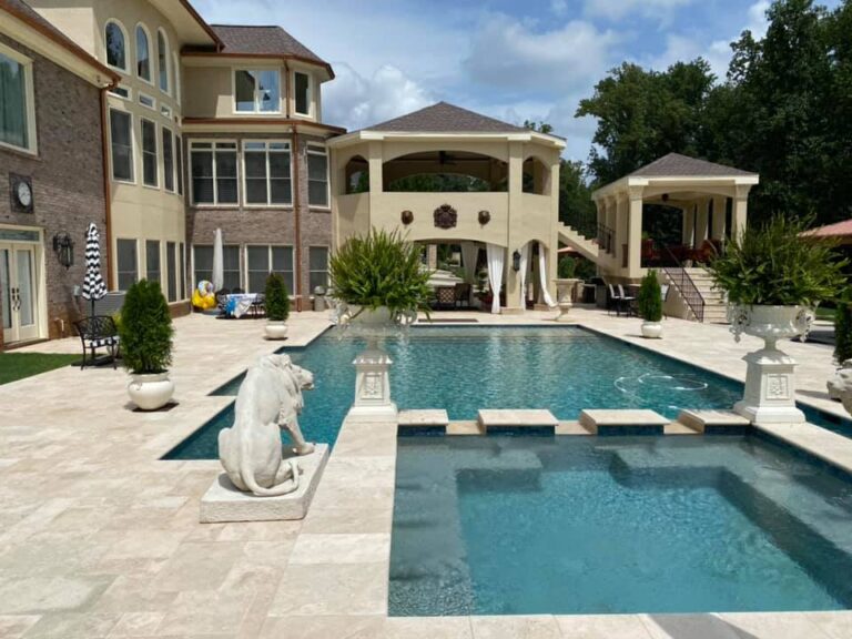 A large backyard with an in-ground pool showcasing the latest pool trends, a hot tub, white statues, patio seating areas, and a multi-story house in the background.