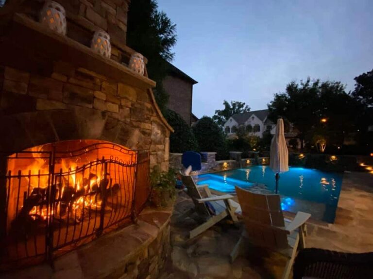 Outdoor stone fireplace with a burning fire next to a lounge area and illuminated swimming pool creates the perfect setting for backyard oasis ideas with pool at dusk.