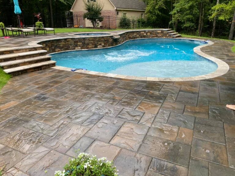 A backyard swimming pool with stone steps and a stone-tiled deck, surrounded by trees and a brick building in the background—ideal for stunning swimming pool renovations.
