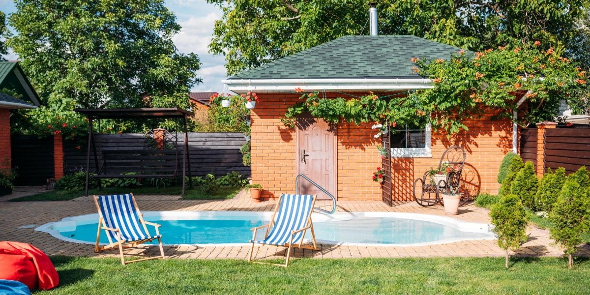 A small backyard inground pool with two striped lounge chairs, a brick pool house, potted plants, and a shaded swing, surrounded by greenery and wooden fences—perfect for relaxing without the high inground pool cost.