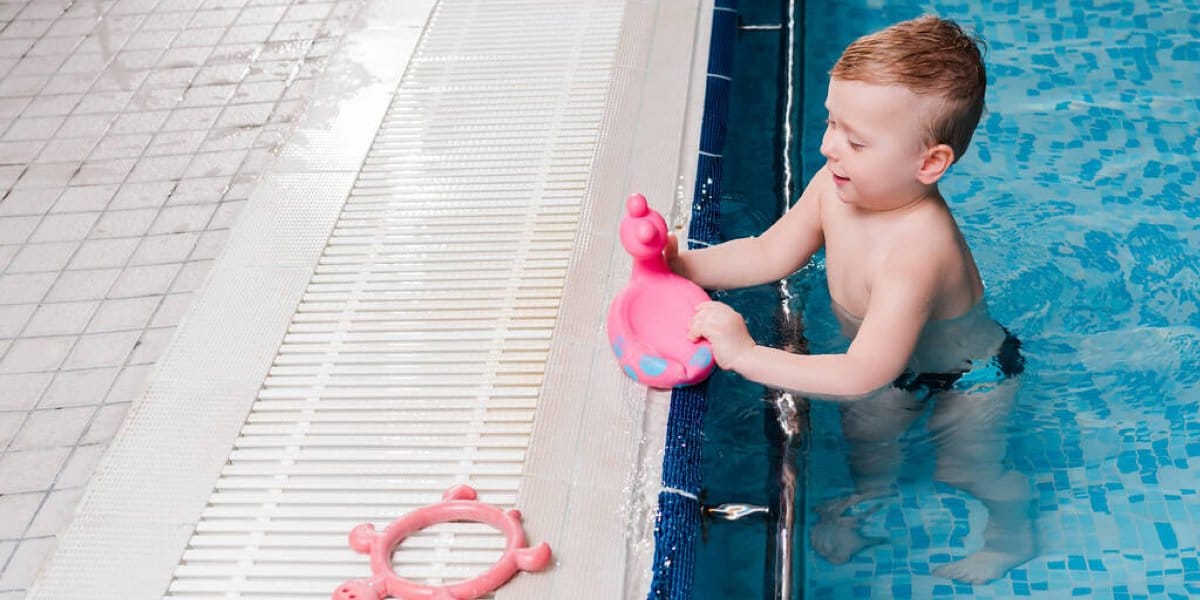 Young boy standing in a swimming pool holds a pink toy while another pink toy, awaiting a wash from those learning how to clean pool toys, rests on the poolside ledge.