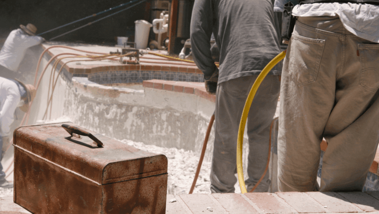 Several workers in protective clothing use equipment to perform gunite pool repair on an empty swimming pool, with a rusted toolbox visible in the foreground.