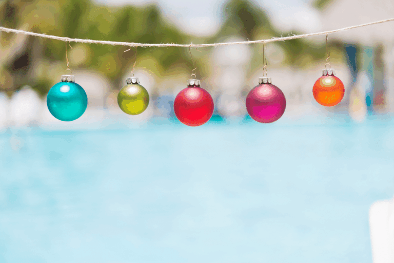 Five colorful Christmas ornaments hang on a string, creating festive christmas pool decorations with a blurred outdoor swimming pool and palm trees in the background.