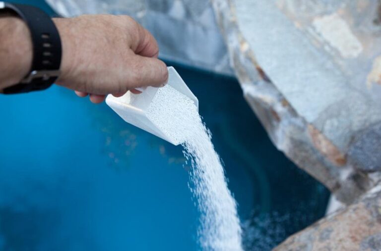 A hand pours granular pool chemical from a white scoop into a blue swimming pool, highlighting the importance of knowing how long after putting chlorine in pool can you swim safely.