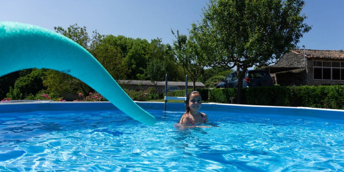 A child swims in an above-ground kids swimming pool with slide outdoors on a sunny day, with a blue pool noodle in the foreground and trees and houses in the background.