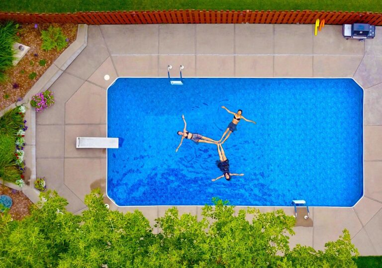 Aerial view of three people floating on their backs in a rectangular outdoor swimming pool, surrounded by an inviting concrete patio and lush greenery—perfect inspiration for those seeking inexpensive pool deck ideas.