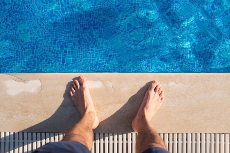 A person stands at the edge of a swimming pool, with bare feet on smooth travertine pool coping next to clear blue water.