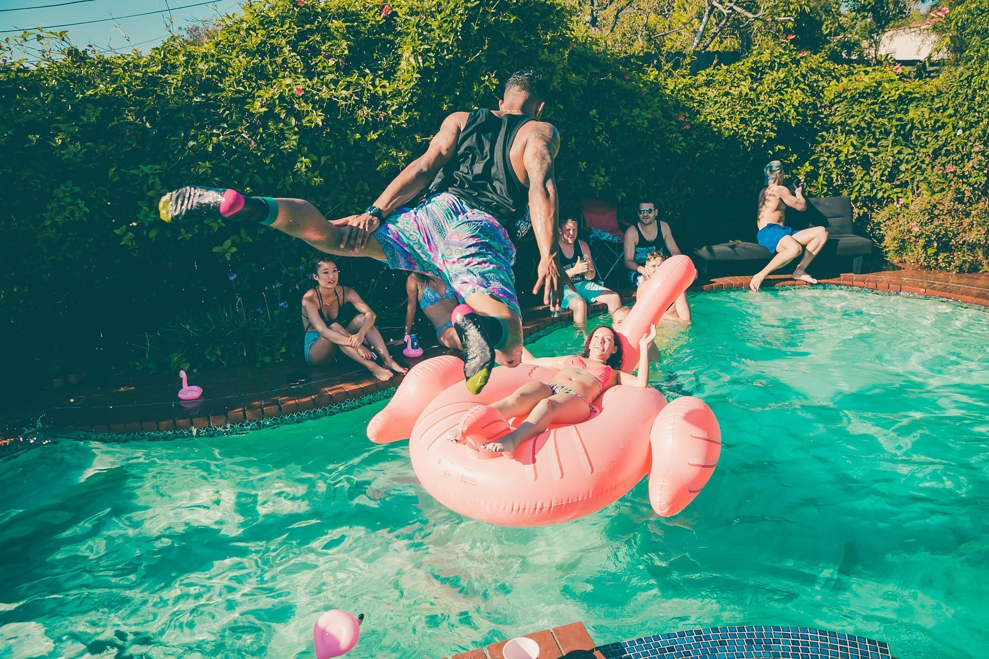 A man jumps into a backyard pool toward a woman on a pink inflatable flamingo, while others watch and relax around the water at a lively Fourth of July pool party on a sunny day.
