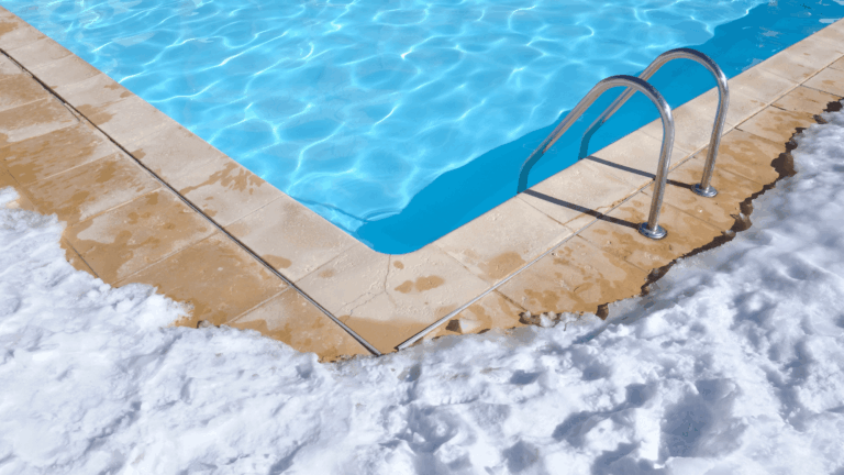 A swimming pool with metal ladder is surrounded by snow and wet tiles, contrasting the blue water with the white snow—a perfect example of why knowing how to winterize a pool is essential.