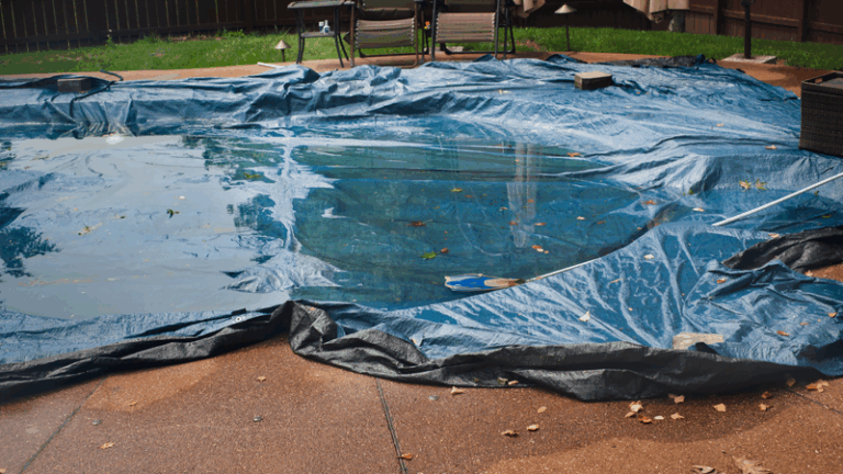 A backyard swimming pool covered with a blue tarp, partly filled with rainwater and scattered leaves, showcases practical swimming pool energy saving tips, surrounded by wet concrete and outdoor furniture.