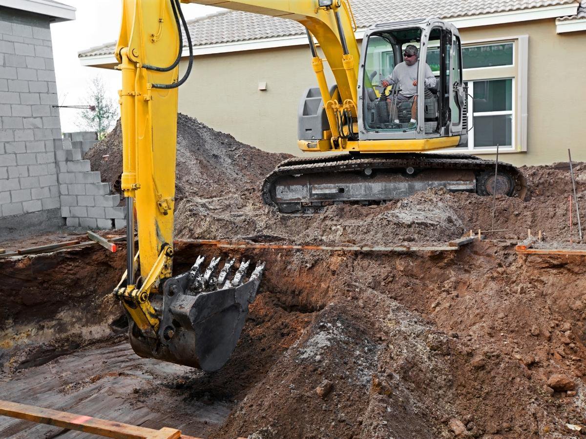 A construction worker operates a yellow excavator, digging a large hole in the ground near a residential building—just like swimming pool contractors preparing for a new installation.