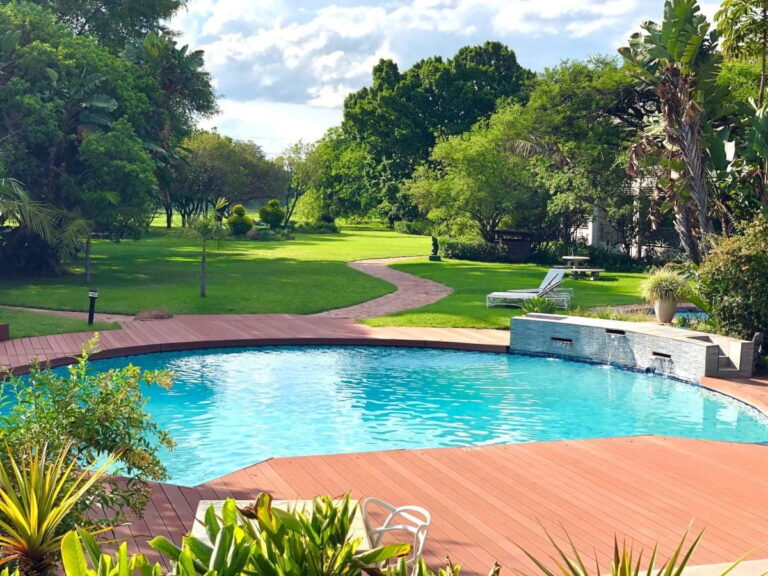 A backyard swimming pool with a wooden deck, surrounded by green grass, trees, lounge chairs, and a stone water feature under a partly cloudy sky—perfect inspiration for pool construction in the fall.