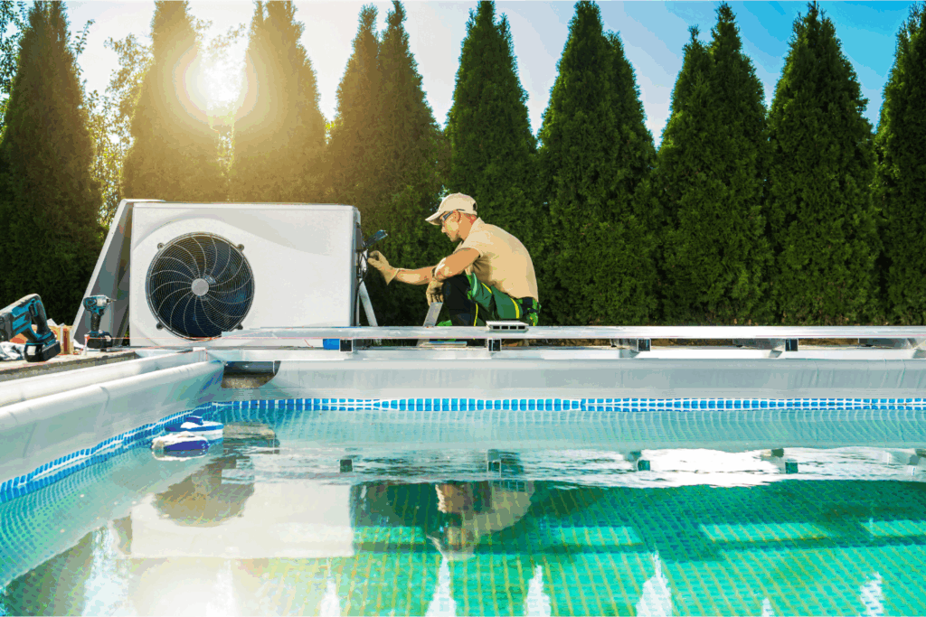 A technician in uniform services a swimming pool heater near an outdoor swimming pool, with tall green hedges in the background.