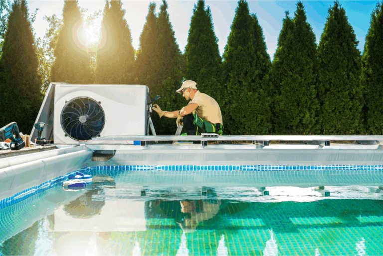 A technician in uniform services a swimming pool heater near an outdoor swimming pool, with tall green hedges in the background.