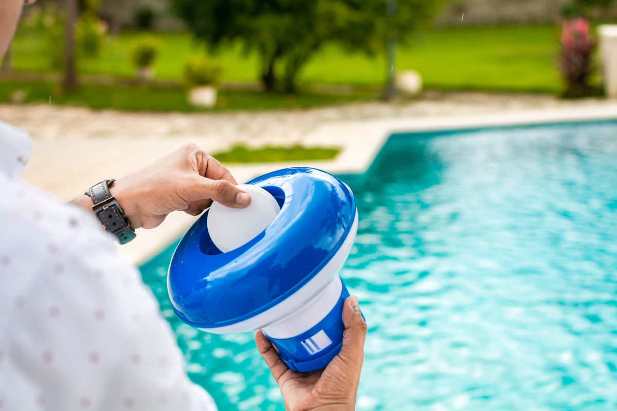 A person practices diy pool maintenance by placing a chlorine tablet into a blue and white floating pool dispenser beside the swimming pool.