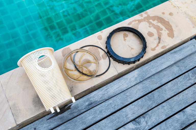 Pool filter parts, including a pool pump basket, rubber gasket, and black ring, are laid out on the edge of a pool beside wooden decking.