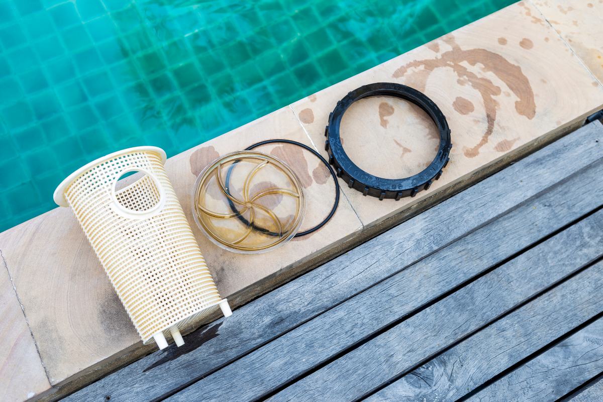 Pool filter parts, including a pool pump basket, rubber gasket, and black ring, are laid out on the edge of a pool beside wooden decking.