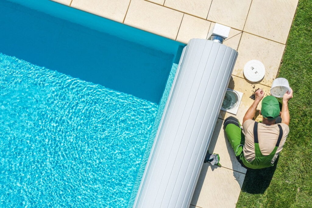 A person in work clothes kneels beside a swimming pool, checking the pool pump and filter with maintenance equipment near the pool's edge on a sunny day.