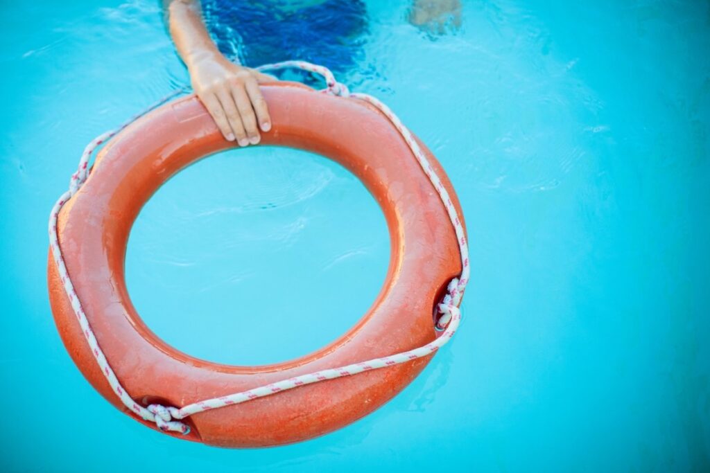 A person’s hand holds onto an orange life preserver floating in a clear blue swimming pool, highlighting the importance of pool safety rules.