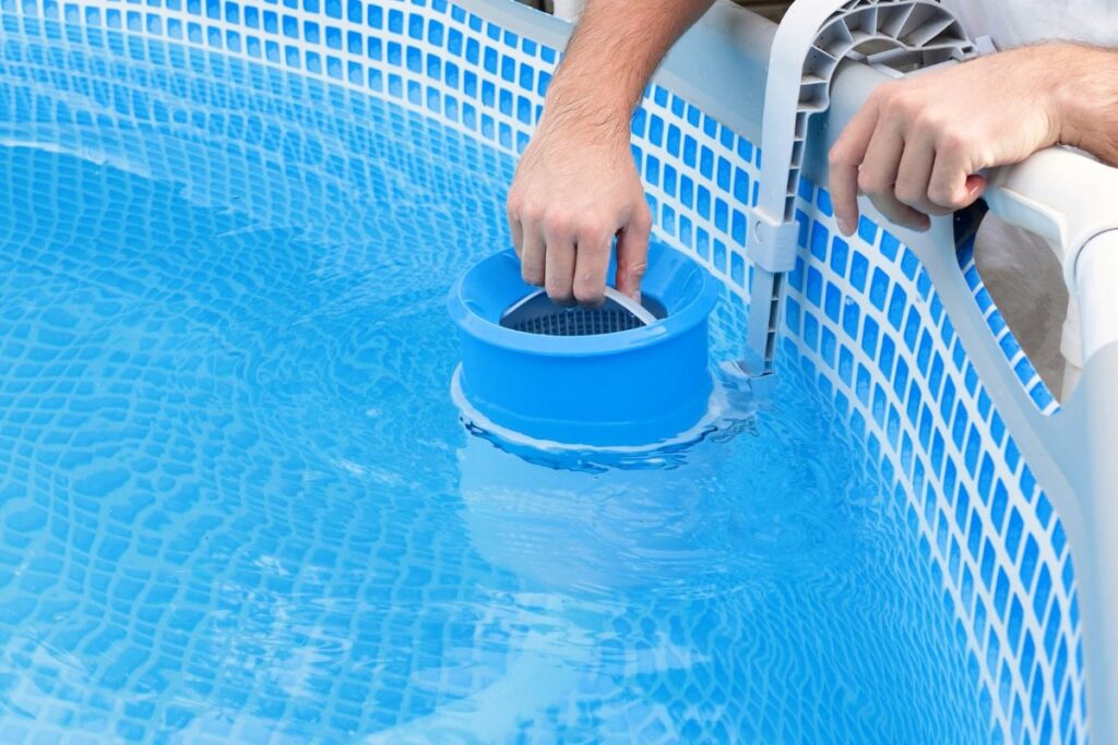 Person installing or adjusting a blue pool skimmer basket inside an above-ground swimming pool with a blue patterned liner, showing how does a pool skimmer work to keep the water clean.