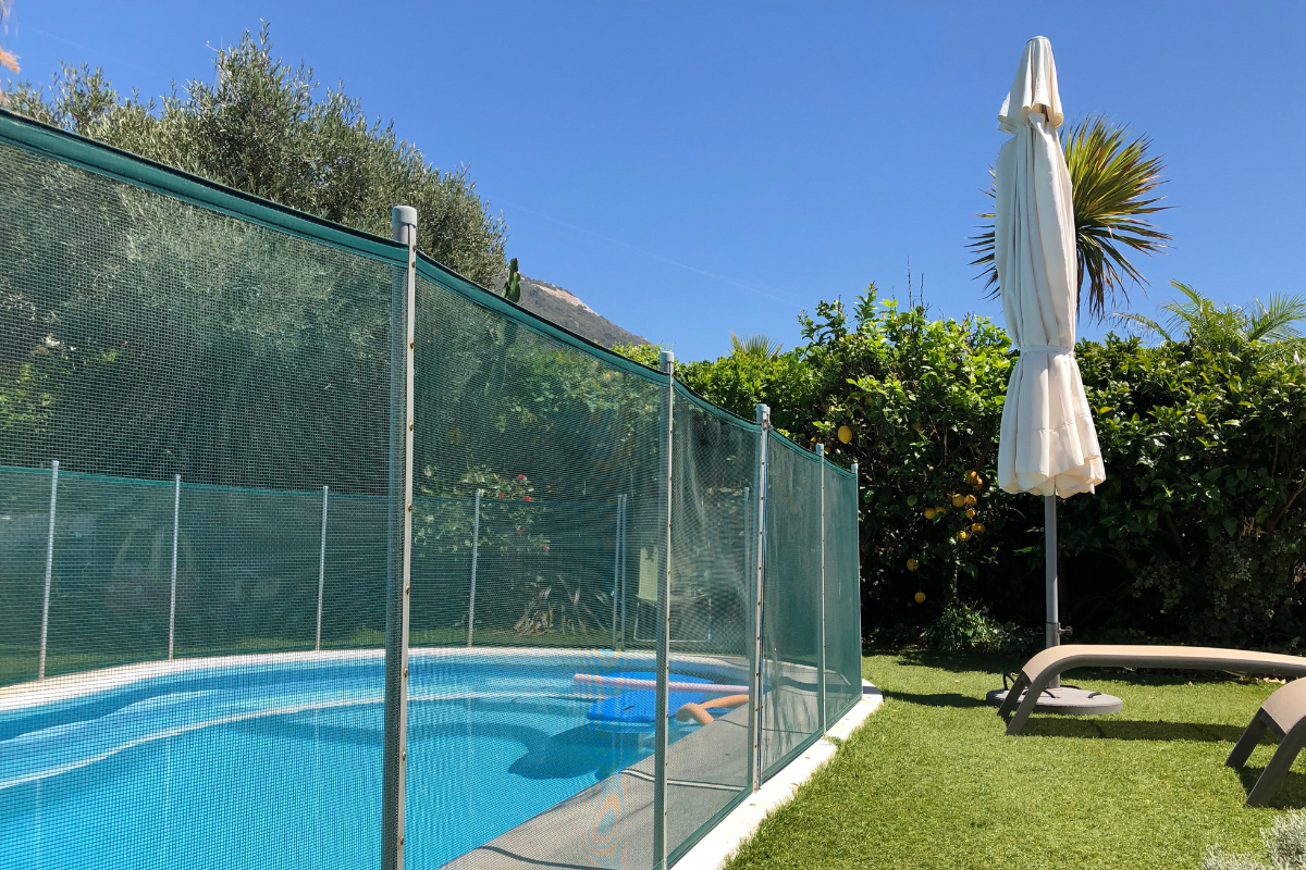 A fenced outdoor swimming pool with a child safety fence sits under a blue sky, surrounded by green plants. A closed white patio umbrella and a lounge chair are on the grass nearby.