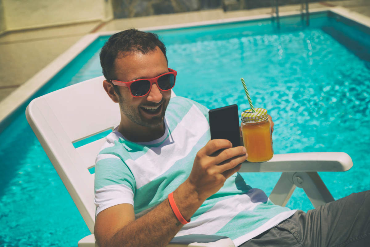 Man wearing sunglasses sits on a lounge chair by a pool, holding a smartphone in one hand—perhaps checking his pool automation app—and a jar of orange juice with a straw in the other.
