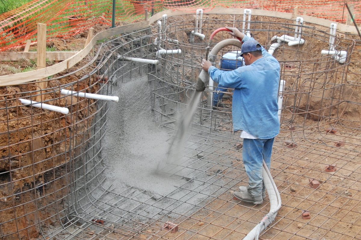 Worker in blue clothing spraying concrete onto steel rebar framework during the construction of an in-ground swimming pool.