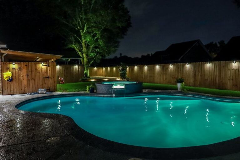 A backyard swimming pool with illuminated blue water at night, inground pool lights, a hot tub, wooden fence, and outdoor lighting along the perimeter.