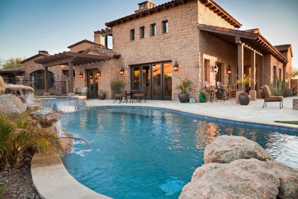 A stone house with large windows and patio seating is shown beside a curved swimming pool with a waterfall feature—perfect inspiration if you're considering installing a swimming pool in your own landscaped backyard.