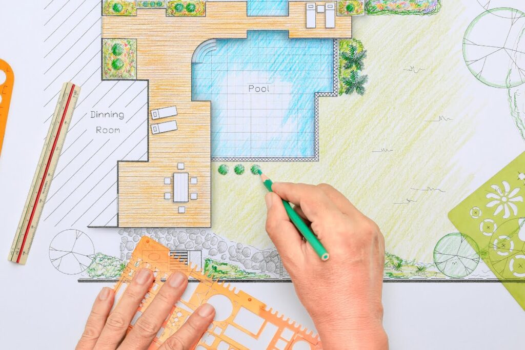 A person draws landscape details on a colored architectural plan of a backyard, referencing a pool planning checklist while adding features like a pool, deck, dining area, and lush greenery using drafting tools.