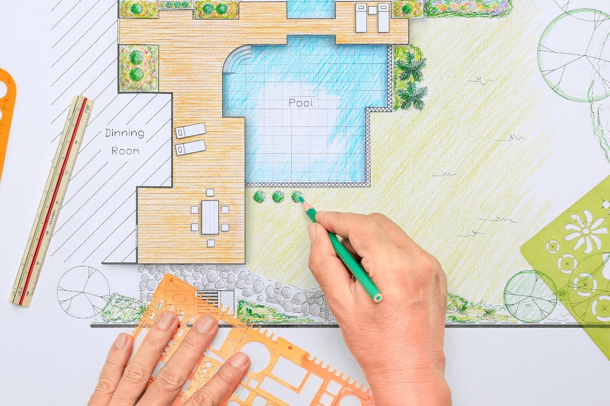 A person draws landscape details on a colored architectural plan of a backyard, referencing a pool planning checklist while adding features like a pool, deck, dining area, and lush greenery using drafting tools.