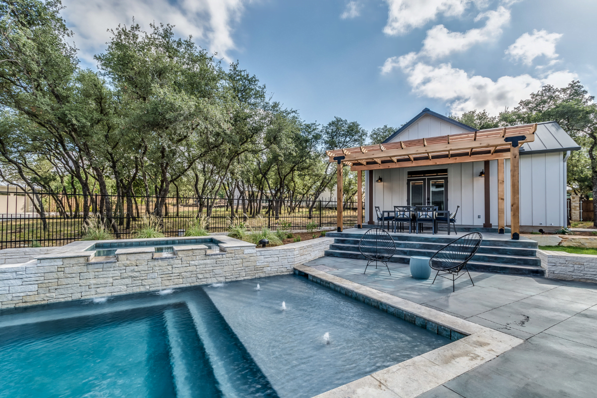 A modern backyard with a custom designed pool, stone wall water feature, patio seating, and a small white guesthouse with a wooden pergola.