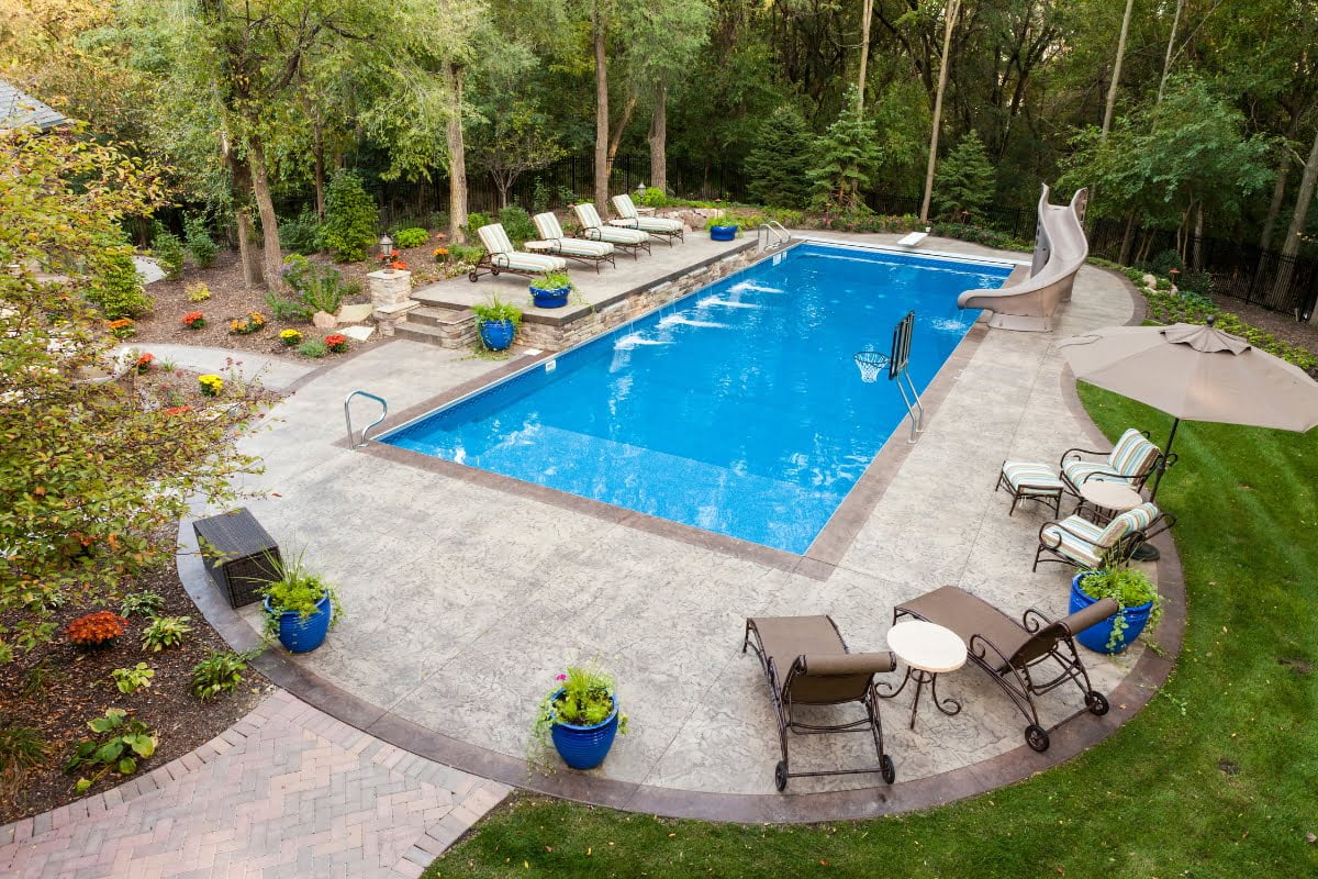 Rectangular pool shape with a water slide, surrounded by lounge chairs, tables, and potted plants; bordered by trees and landscaped greenery.