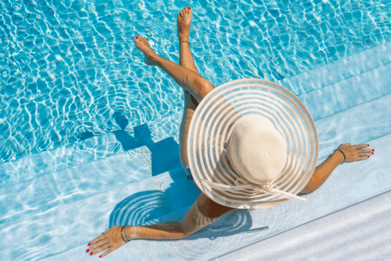 Person wearing a wide-brimmed sun hat sits on pool steps with feet in the water, enjoying the benefits of a salt water pool as gentle ripples and shadow patterns dance across the surface.