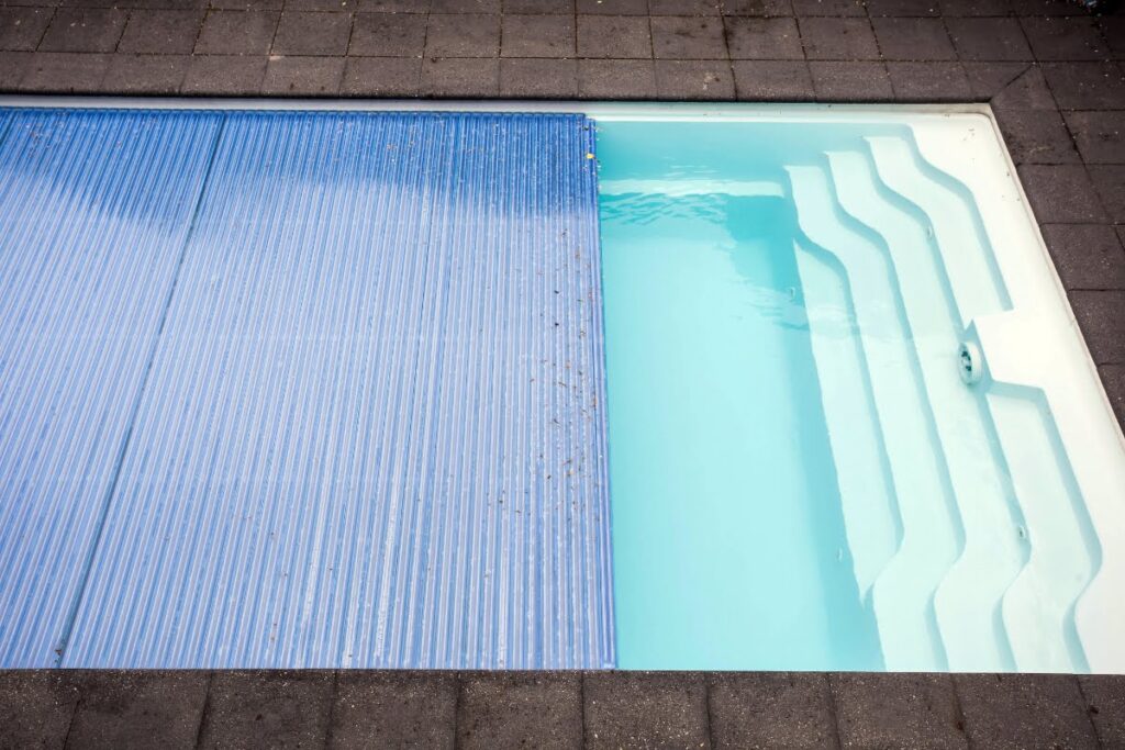 A rectangular swimming pool with white steps, partially covered by a blue slatted pool cover, surrounded by gray paving stones—perfect for closing the pool for winter.