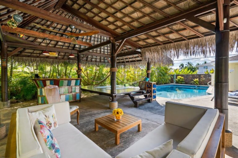 Covered outdoor lounge area with cushioned seating, a wooden coffee table, a hammock, and a pool table beside the swimming pool, this inviting pool cabana is surrounded by lush tropical plants.