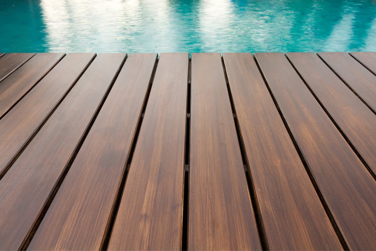 Close-up view of a wooden pool deck material with dark brown planks next to a body of blue water, likely a swimming pool.