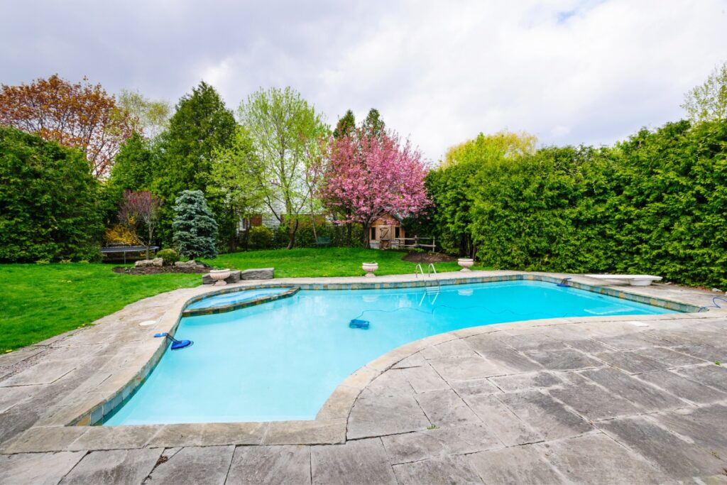 A backyard with a pool surrounded by trees and shrubs. The pool deck material is a stunning addition to the overall landscape.