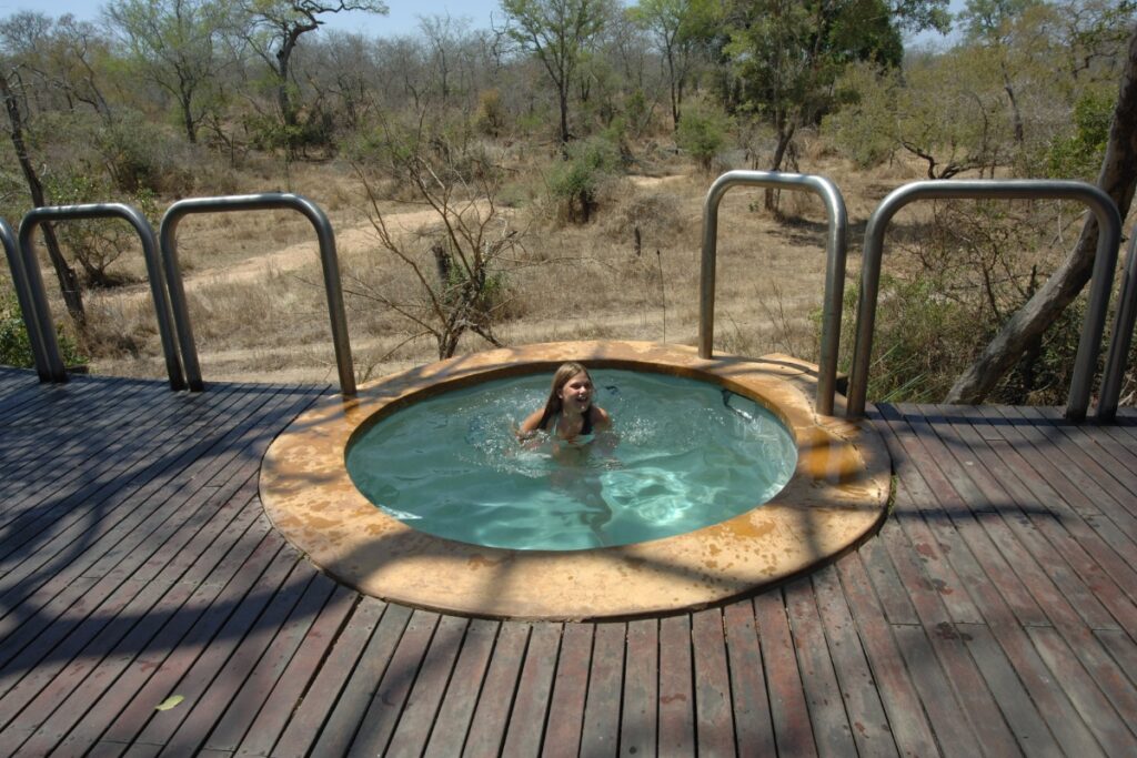 A woman is relaxing in a hot tub on a wooden deck next to an inground plunge pool.
