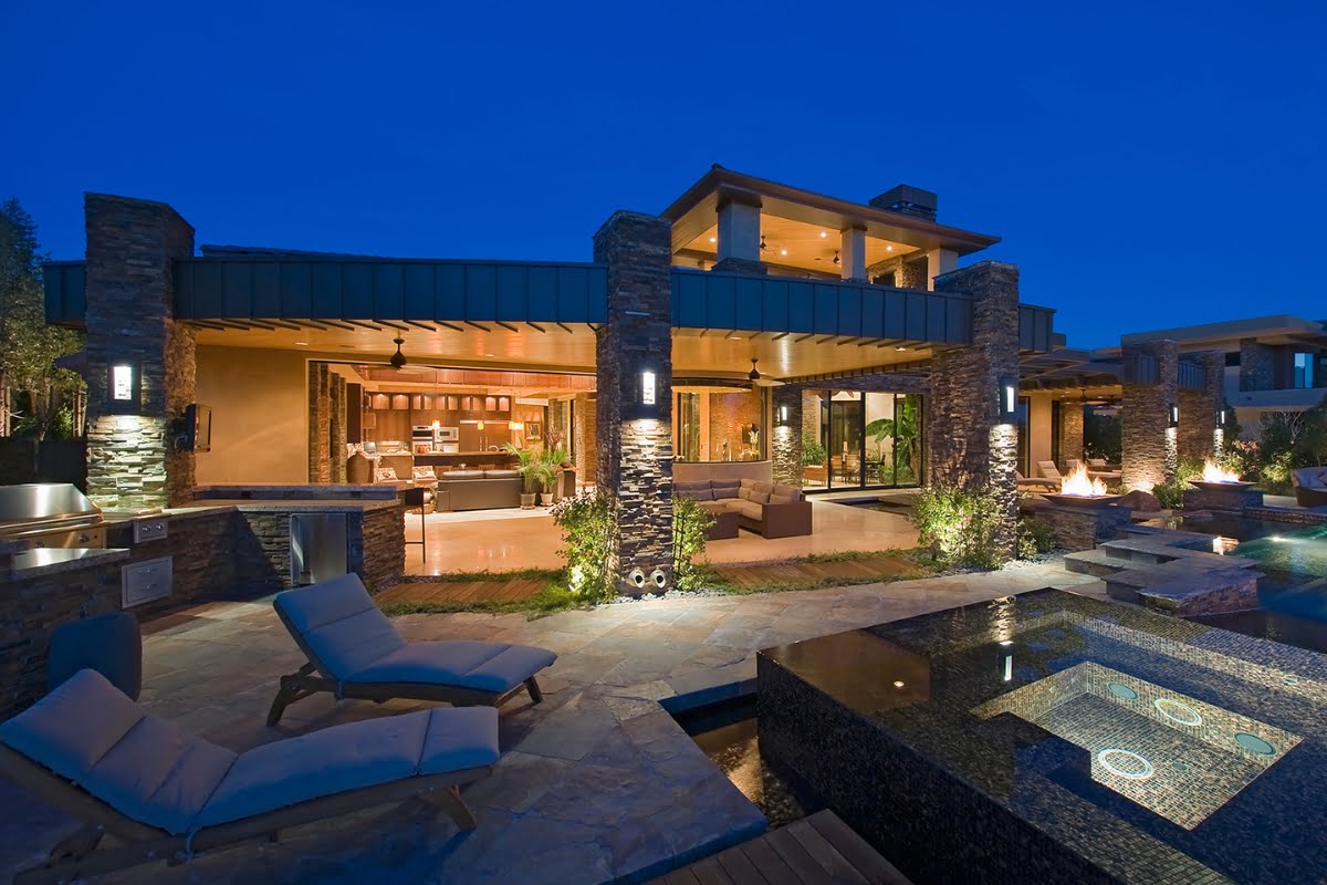 Modern luxury house at dusk with large glass windows, outdoor seating, pool, hot tub, inground plunge pools, lounge chairs, and illuminated stone pillars.