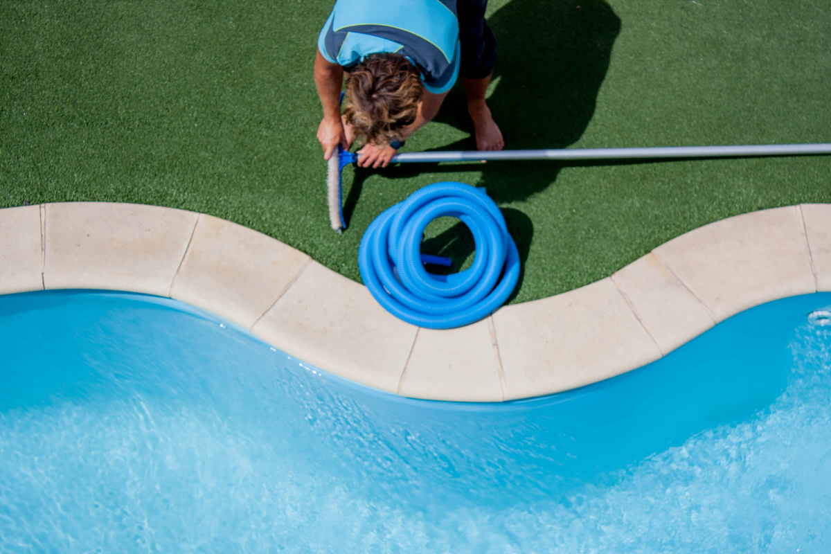 A person stands on artificial grass by a swimming pool, holding a vacuum pole and preparing a blue hose for eco friendly pool maintenance.