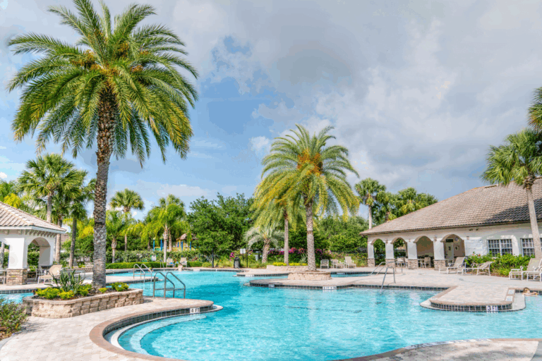 Outdoor swimming pool featuring the latest pool design trends, surrounded by palm trees, lounge chairs, and two pavilion-style buildings under a partly cloudy sky.