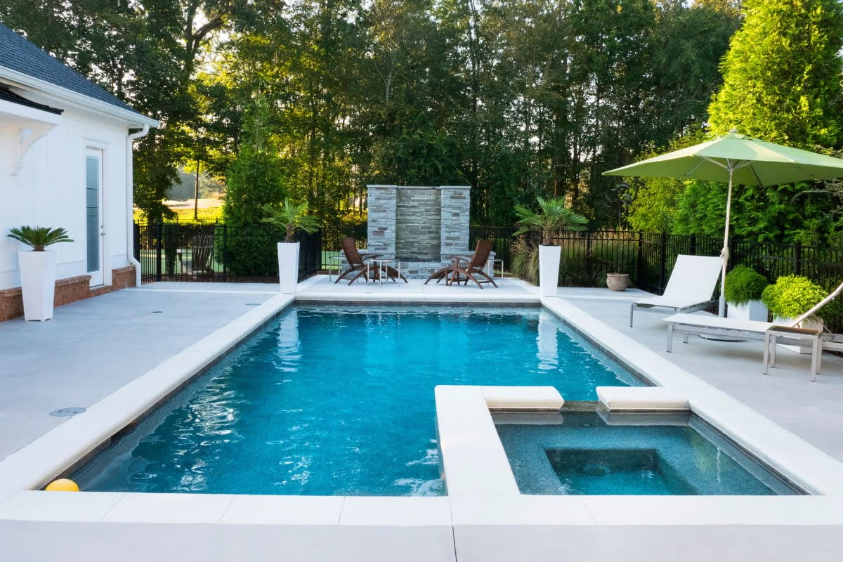 Rectangular backyard swimming pool with attached hot tub, surrounded by lounge chairs, an umbrella, potted plants, and a seating area with a small stone wall at the far end—perfect for those seeking energy efficient pools.